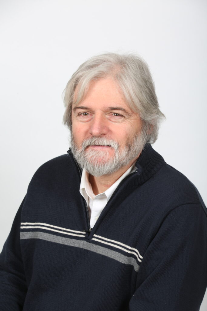 A headshot of Dr. Keith Thompson. He is smiling and has medium length grey hair and a grey beard. He is in a dark blue sweater.